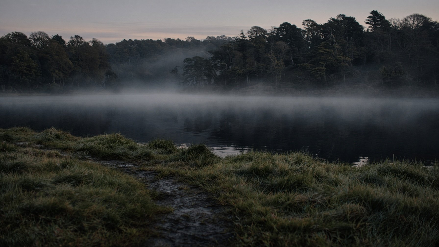 First light and mist over a still Cornish lake, empty bank in the foreground